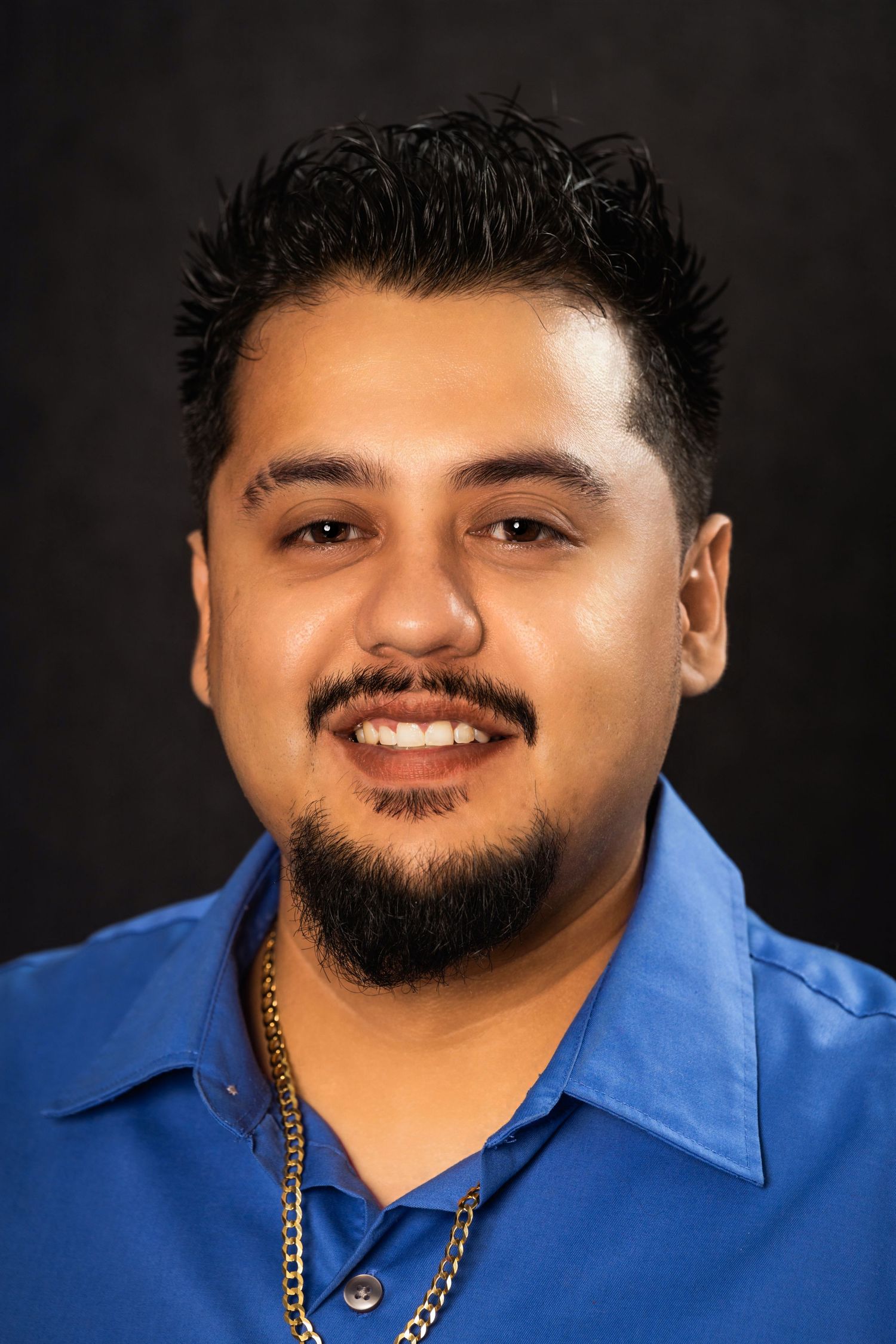 Studio portrait of a person in blue shirt with gold chain against dark background.