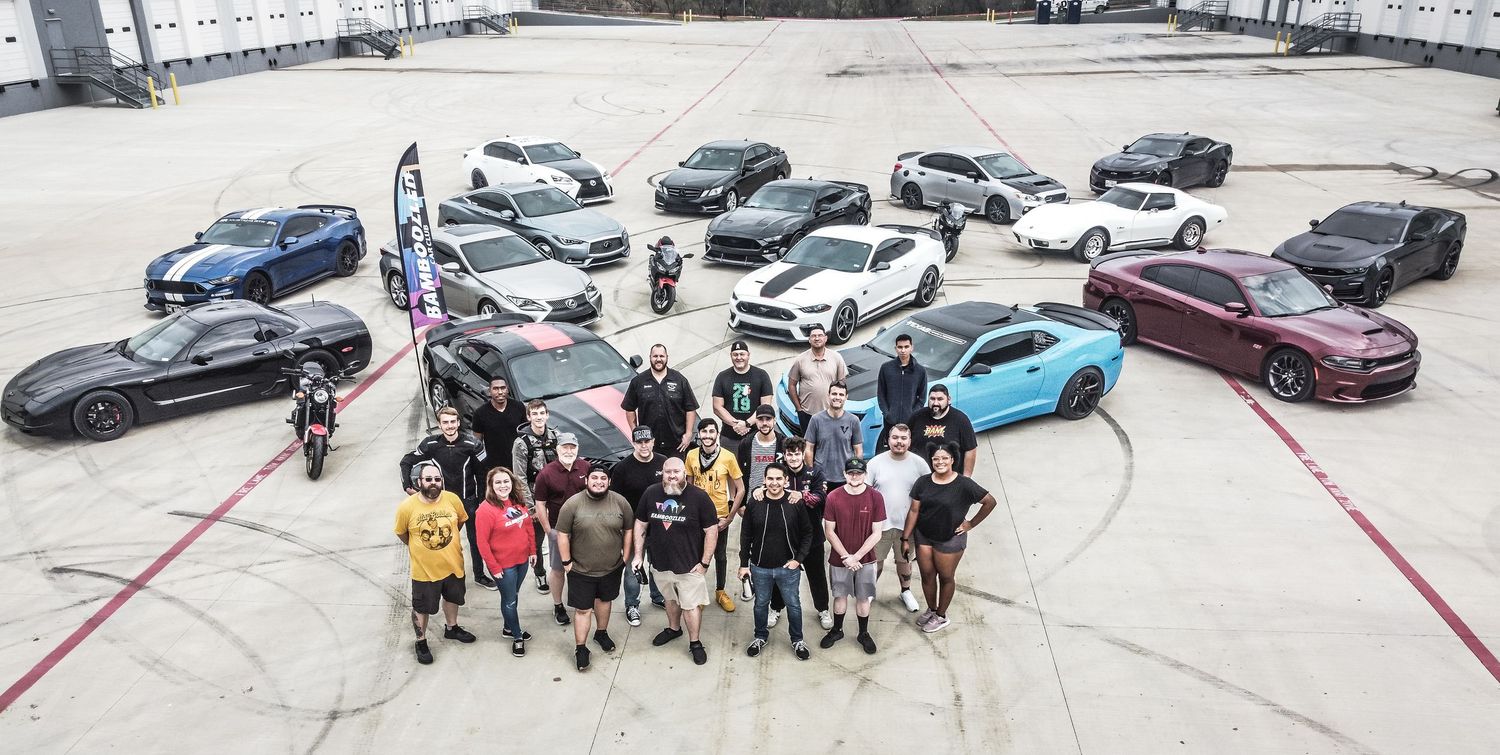 Group gathered around collection of sports cars in industrial parking area.