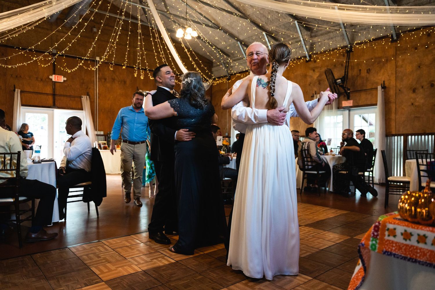 Wedding guests dance together under warm string lights and fabric draped ceiling at a rustic reception venue.