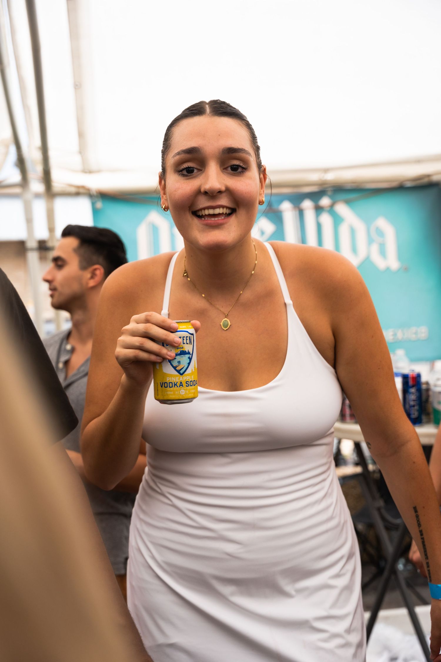 Person in white tank top holding beverage can at outdoor summer event.