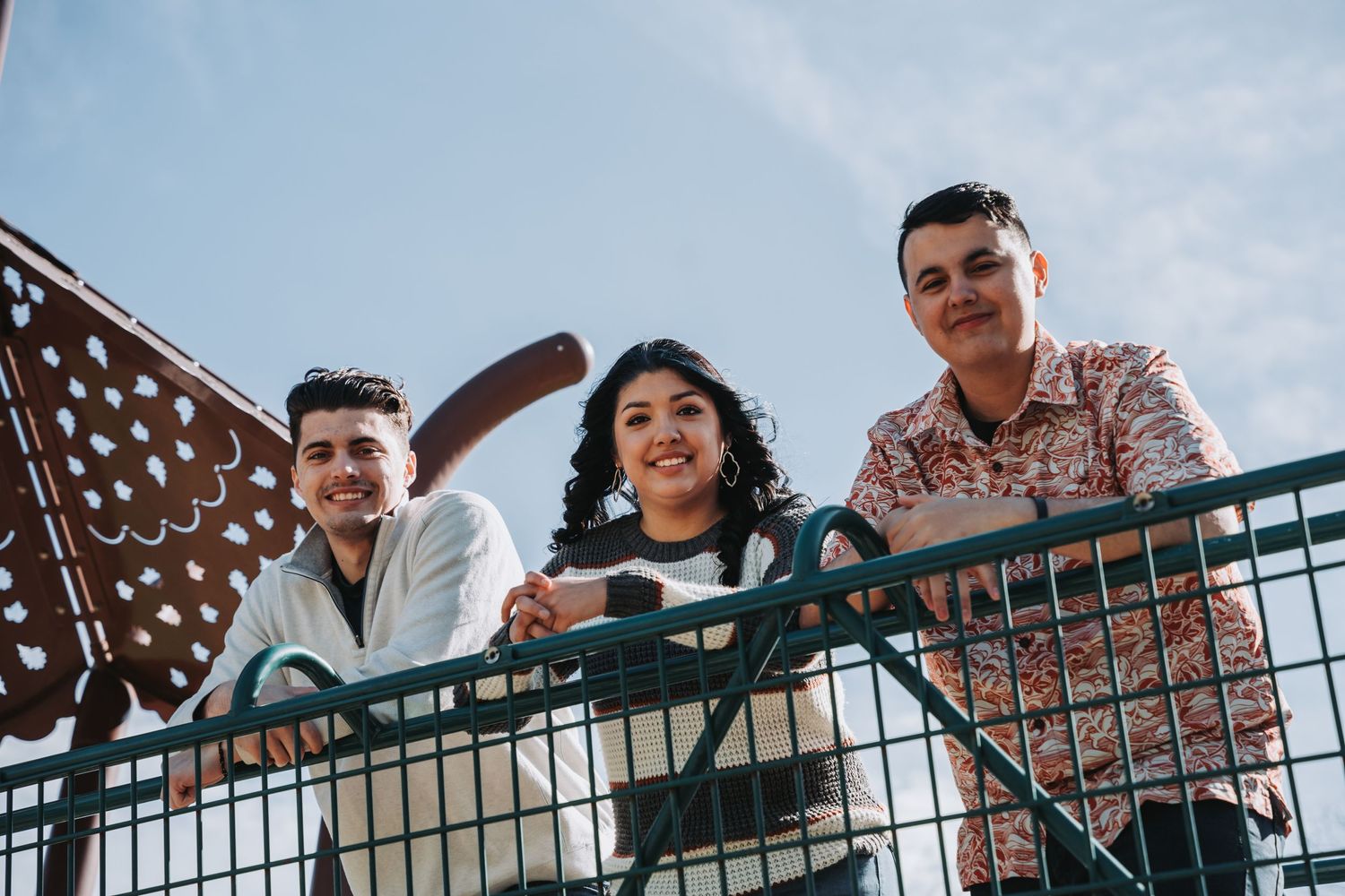 Three friends smile and lean against a metal railing outdoors against a bright sky with decorative wings in background.