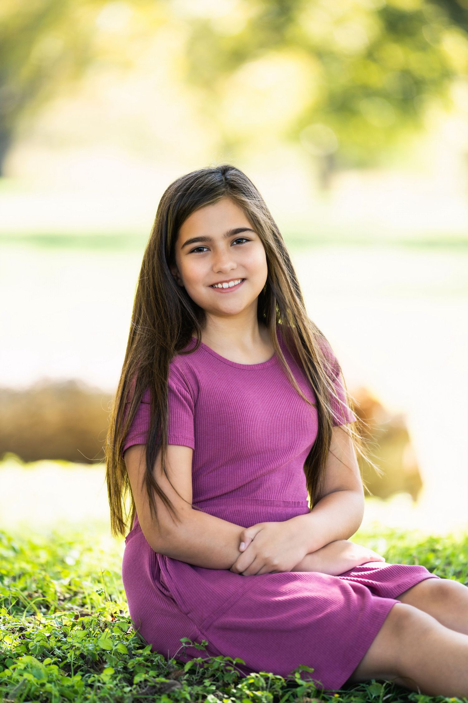 Young person in a purple ribbed dress sits smiling on grass in a sunny outdoor setting with bokeh background.