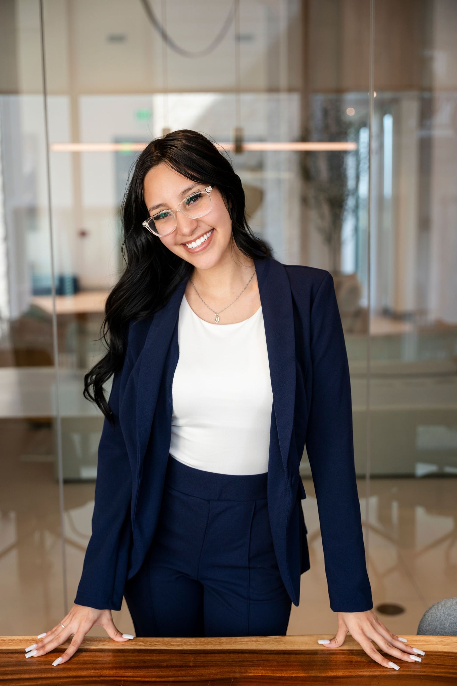 Professional in navy business suit standing confidently at wooden desk in modern office with glass walls.
