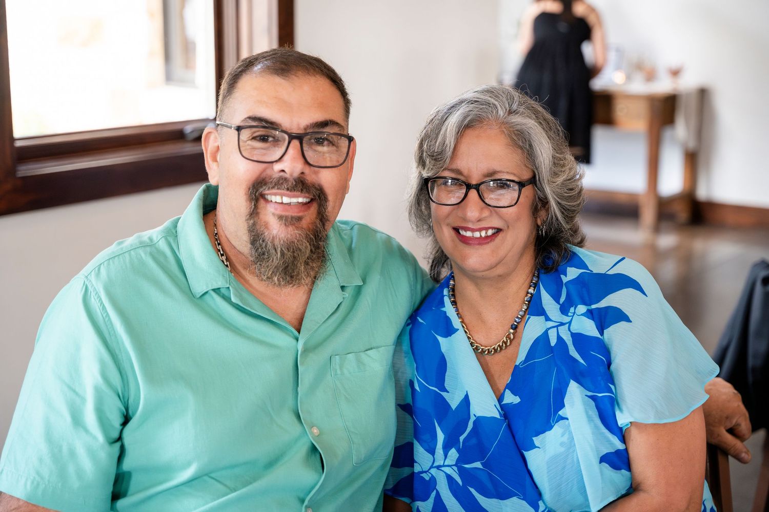 Two people wearing glasses sit close together and smile warmly at the camera in casual attire.