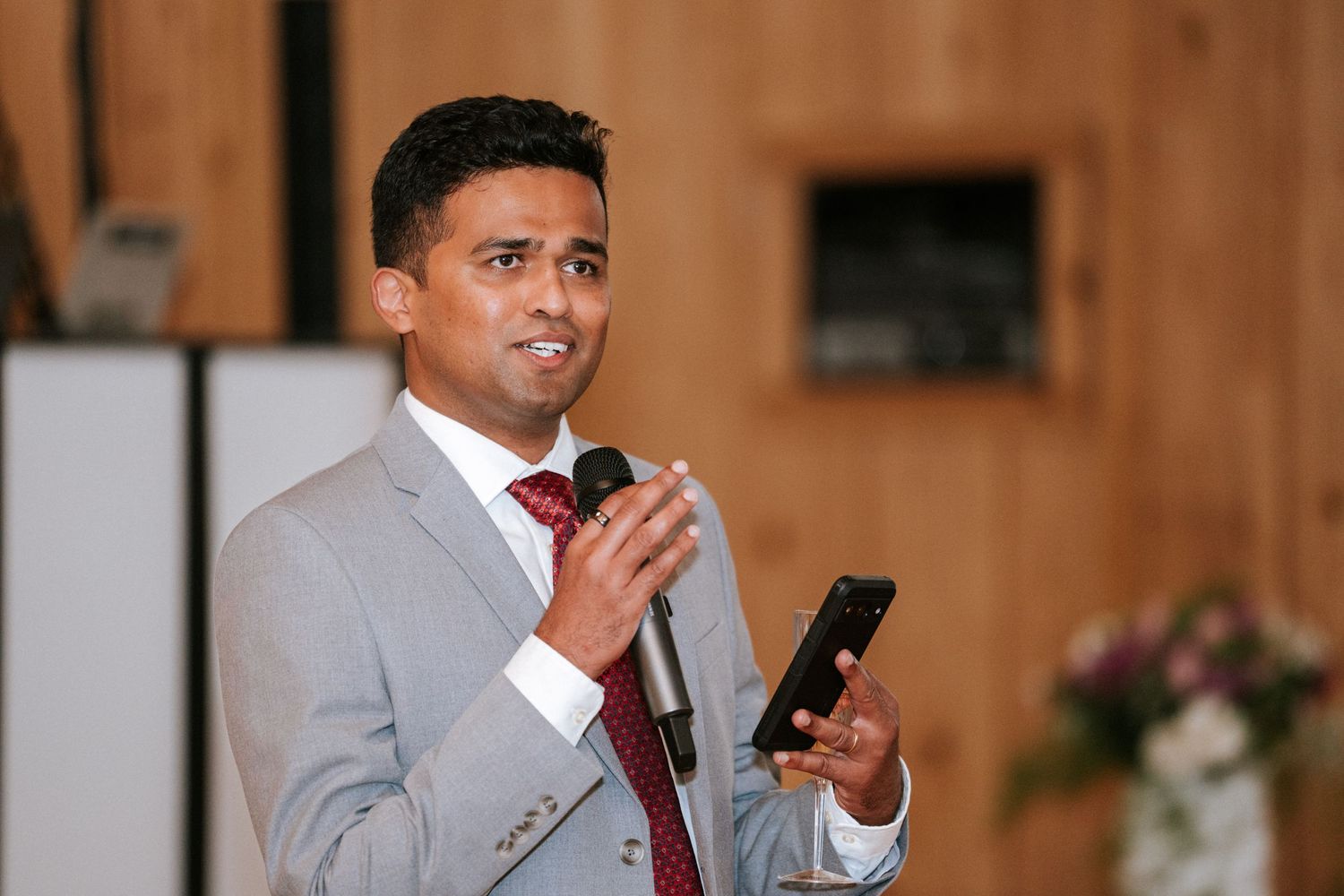 A speaker in a gray suit and red tie addresses guests at a formal event.