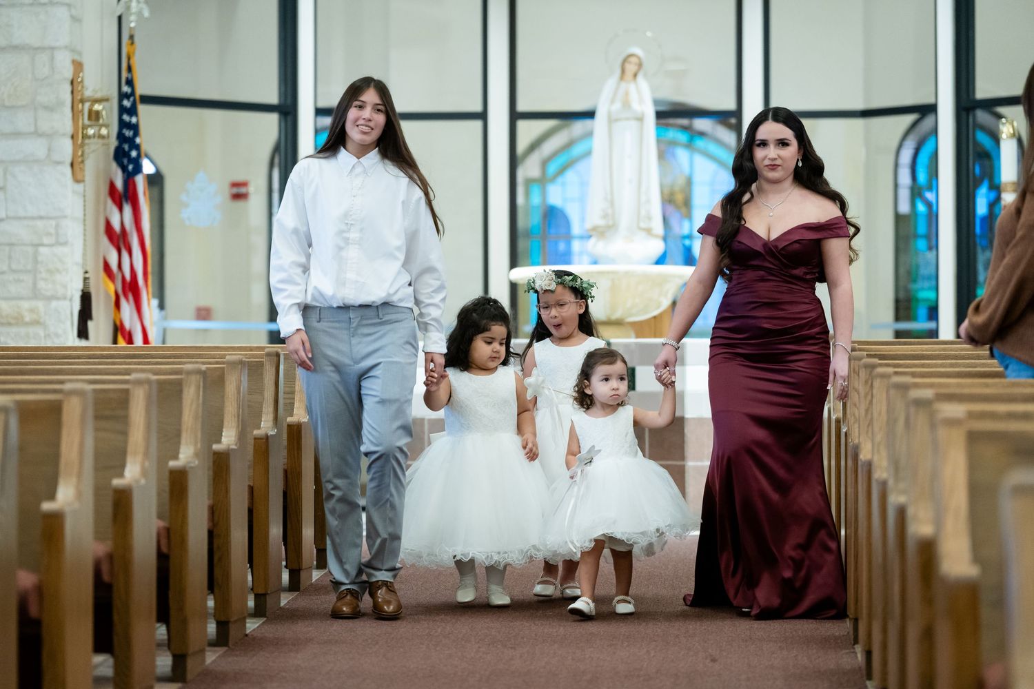 Family walking down church aisle with two flower girls in white dresses and woman in burgundy gown.