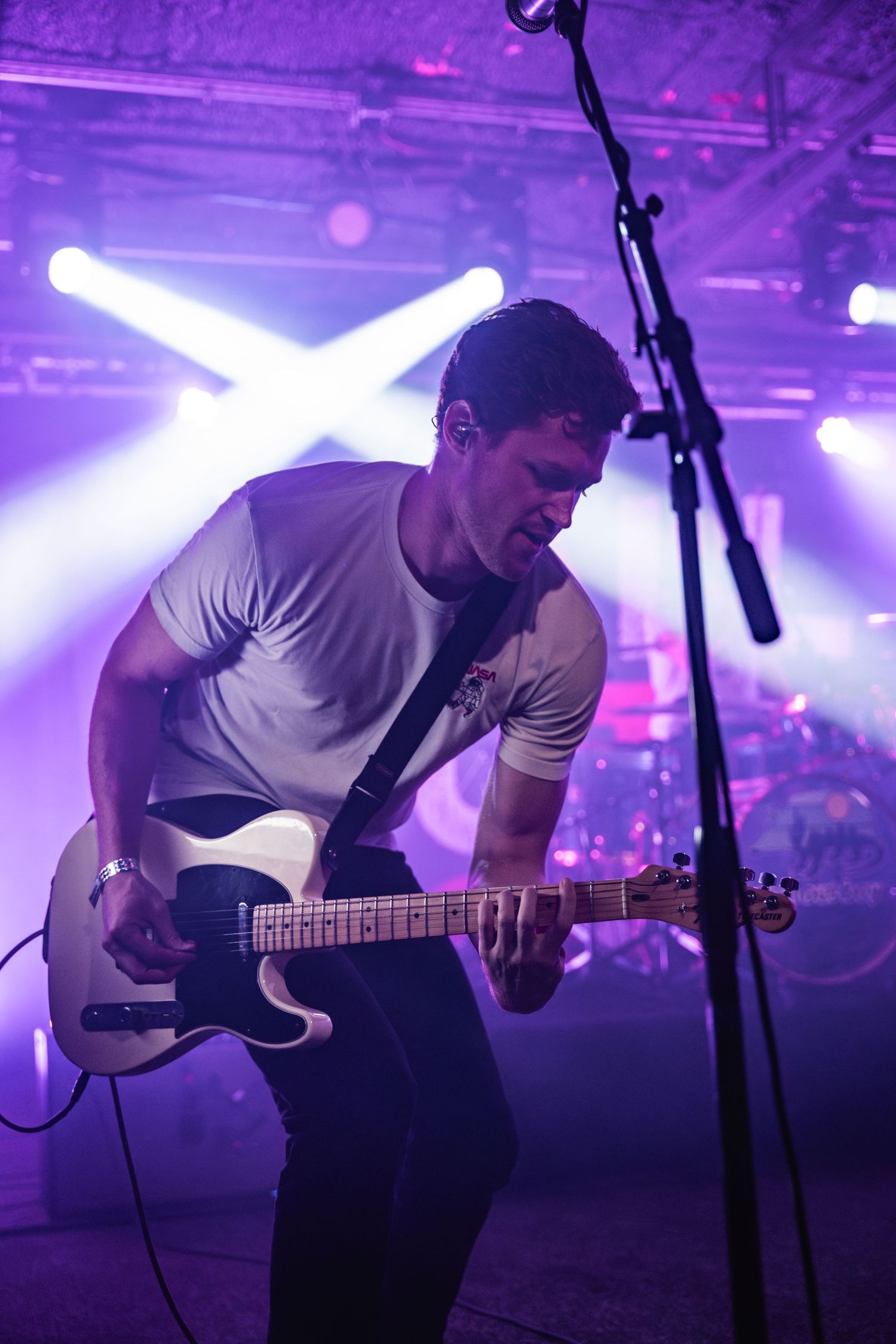 Musician performs on stage with electric guitar under dramatic purple concert lighting and crossed spotlights in background.