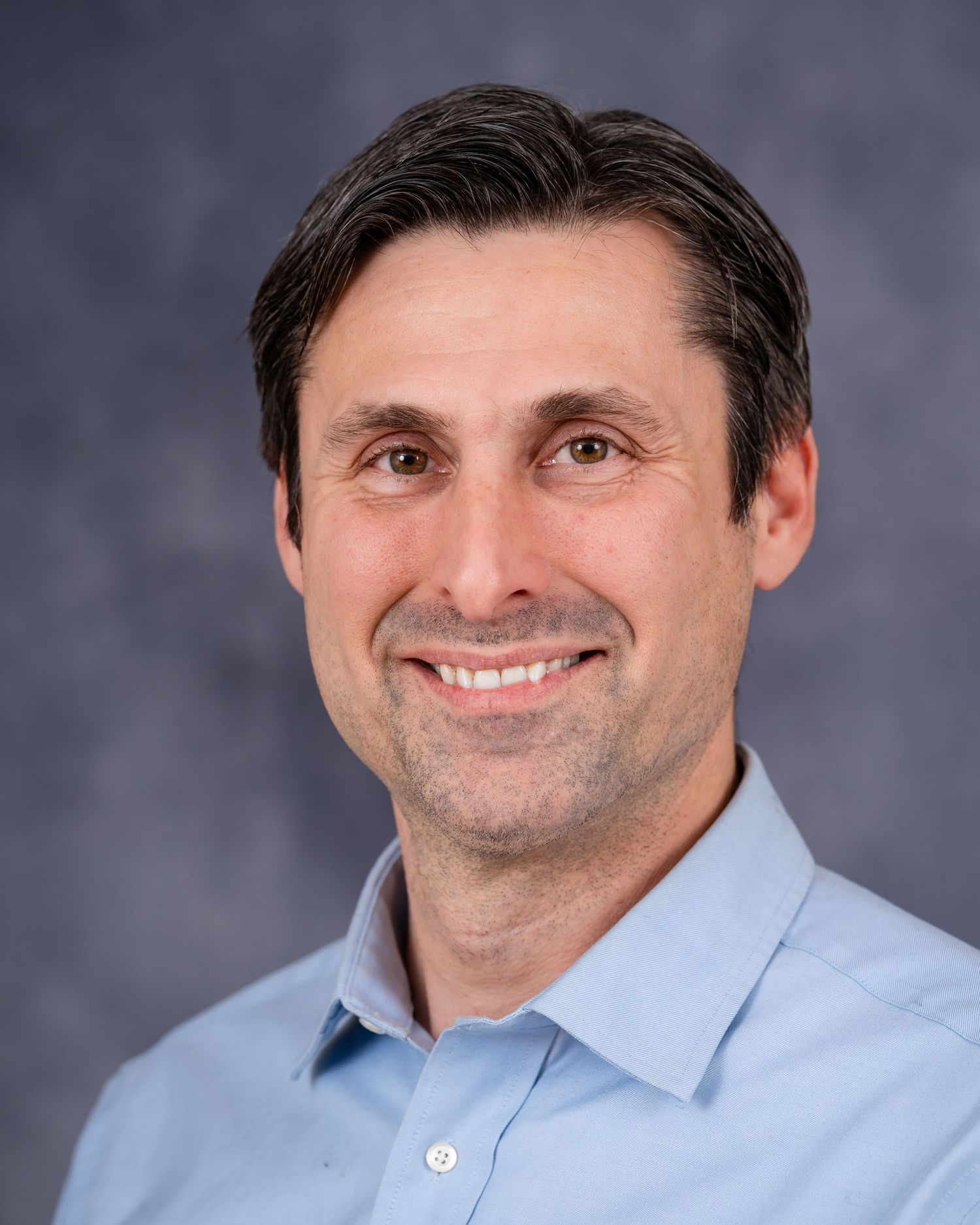 Professional headshot of a person in a light blue button-down shirt against a gray background.