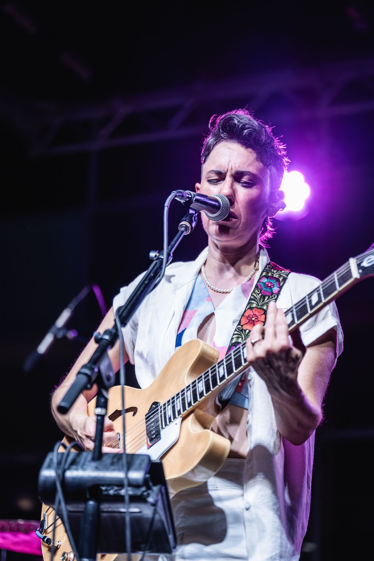 Musician playing acoustic guitar and singing into microphone under purple stage lighting.