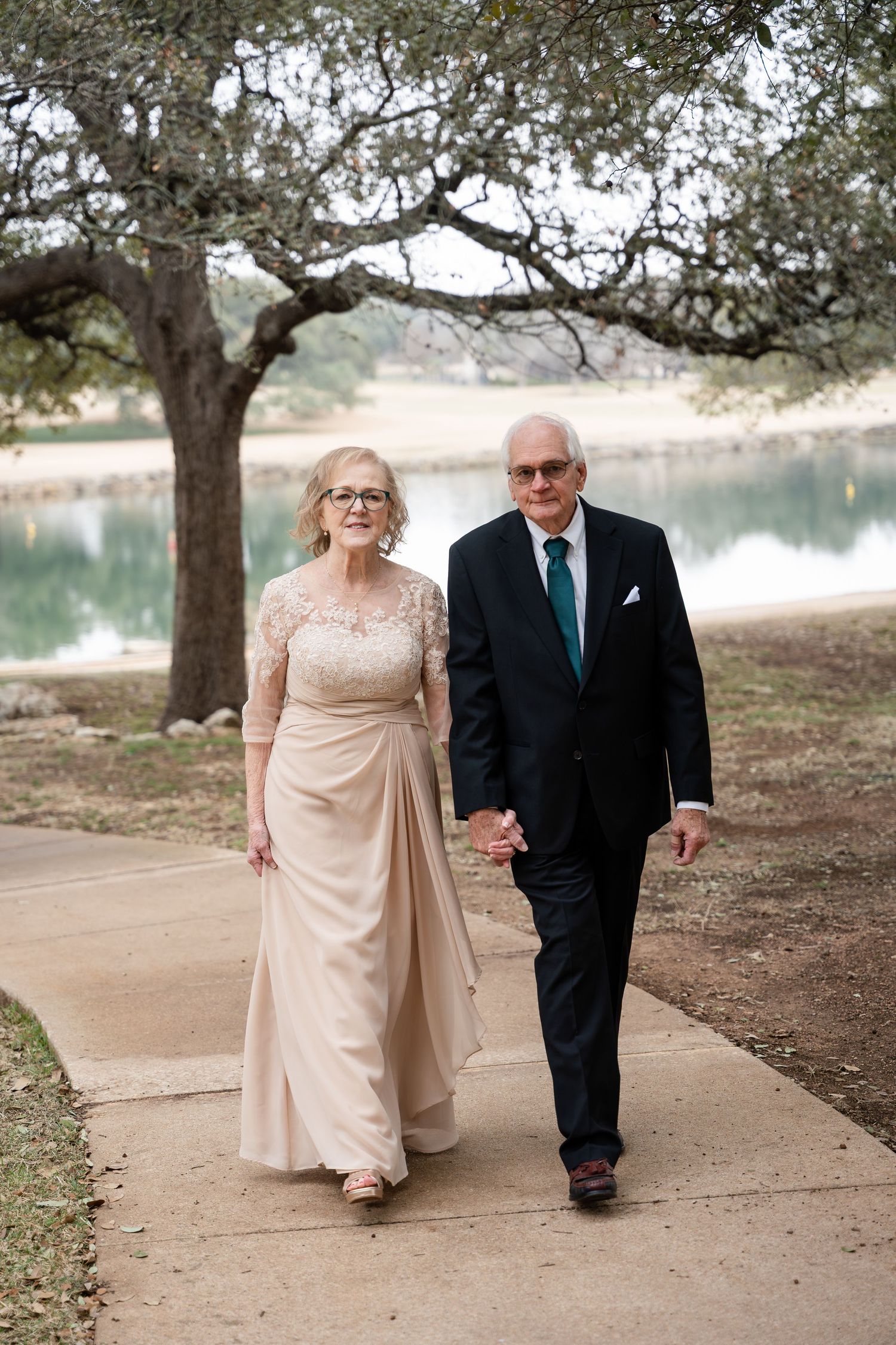 An older couple holds hands while walking along a lakeside path under a large oak tree.