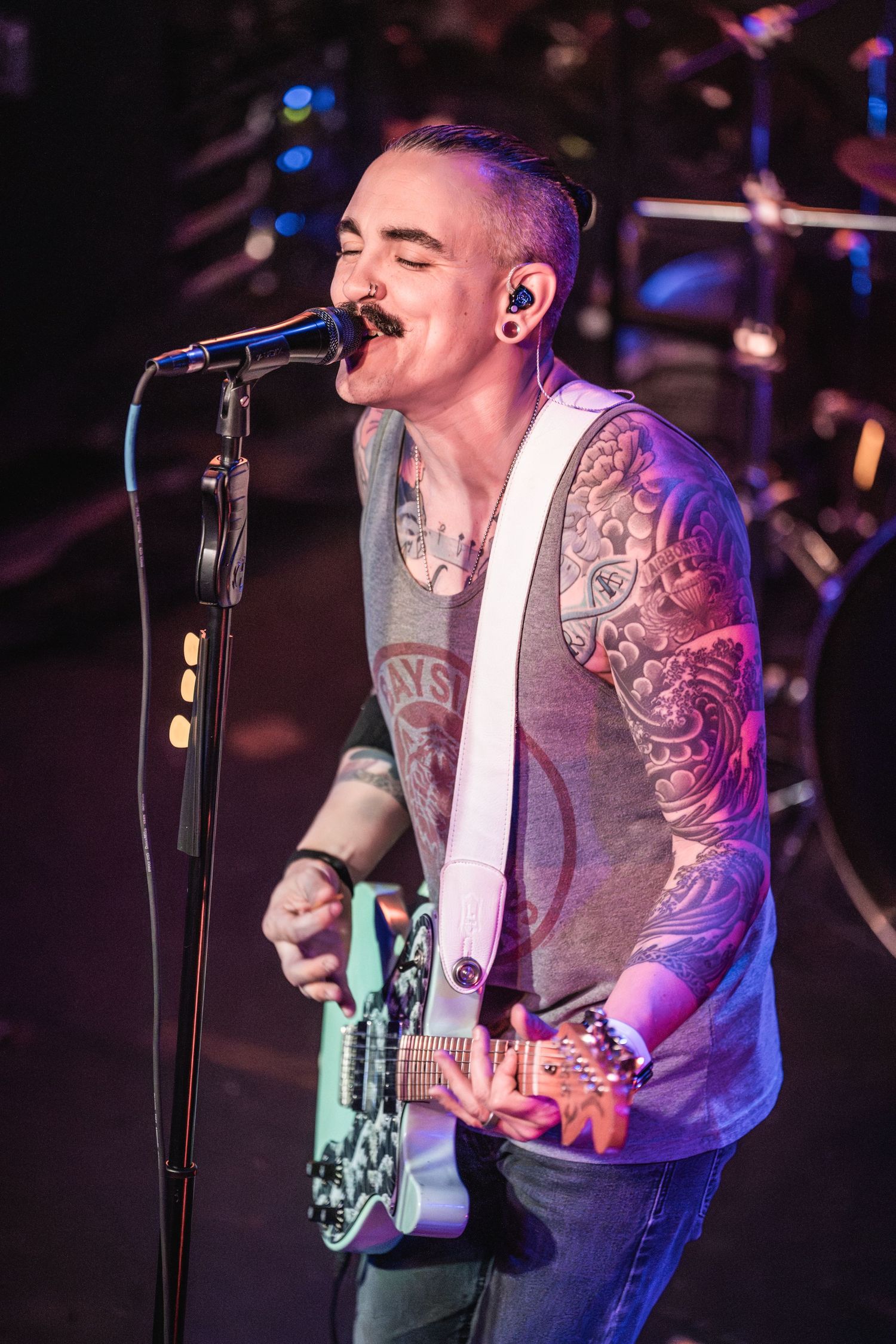 A tattooed musician sings into a microphone while playing guitar on a dimly lit stage with purple lighting.
