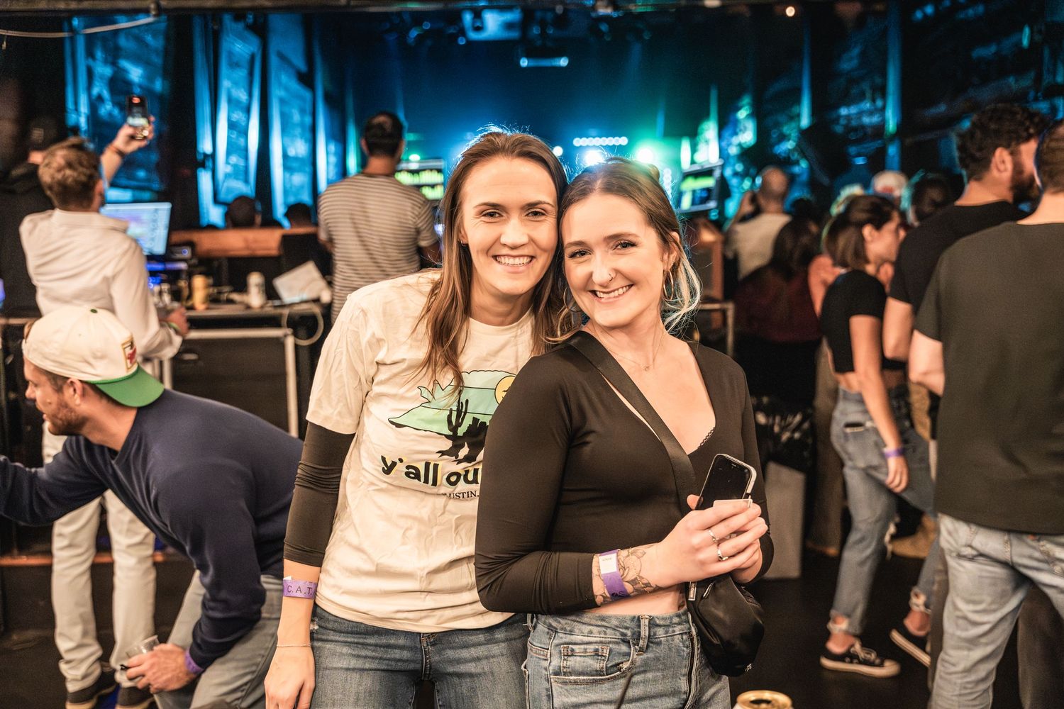 Two concert attendees pose together at music venue with crowd in background.