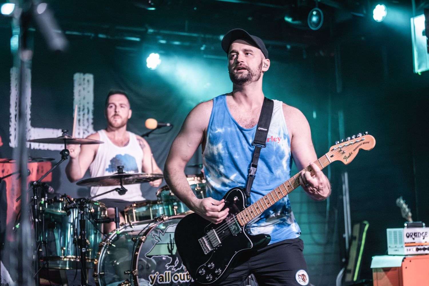 Musicians playing drums and guitar perform energetically on stage with blue lighting.