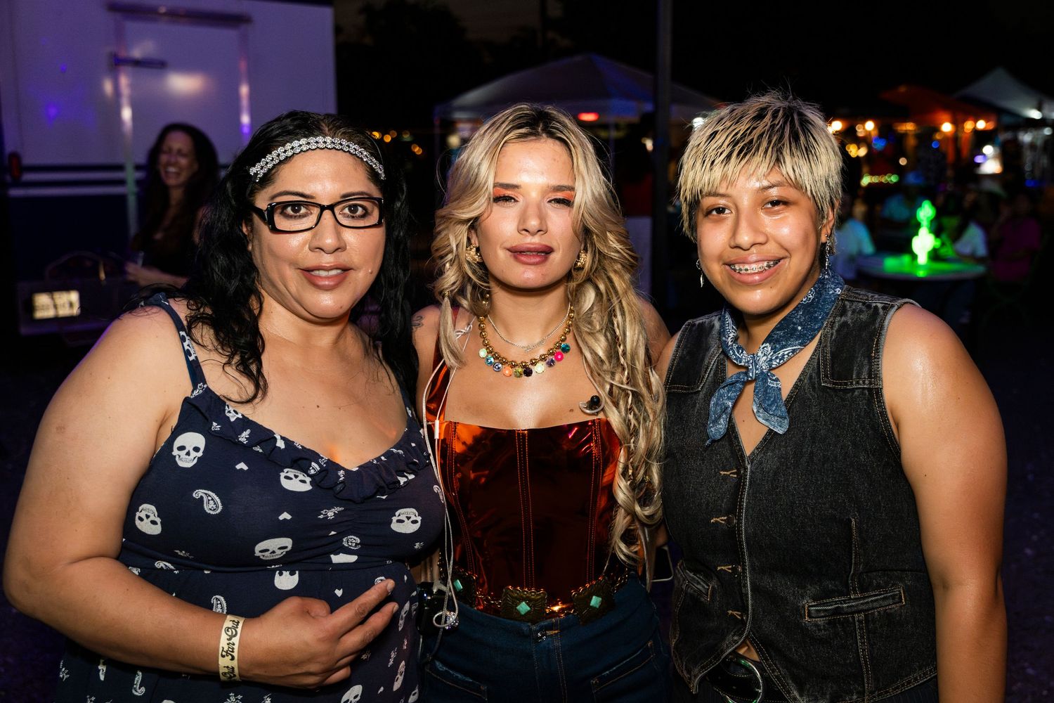 Three friends pose together at a nighttime social event with colorful lighting in the background.