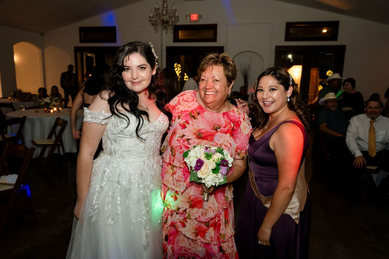 Three people pose together at an evening reception with a bouquet of flowers and colorful party lighting.