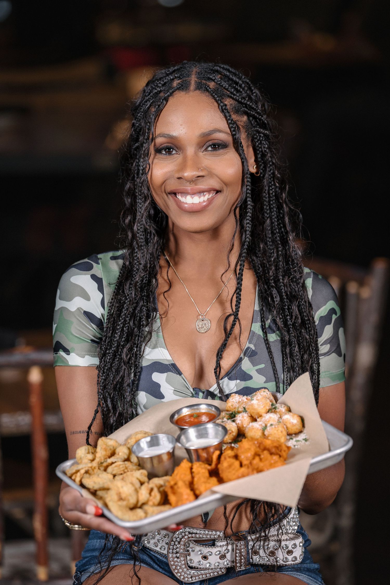 Server in camo crop top presents platter of appetizers with bright smile.