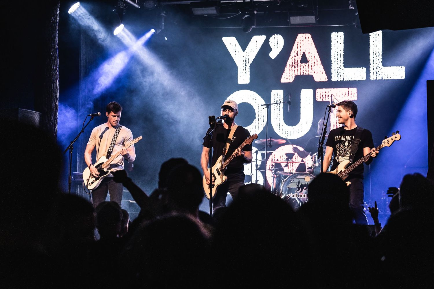 Rock band playing energetic performance under large Y'ALL OUT sign with dramatic blue lighting.