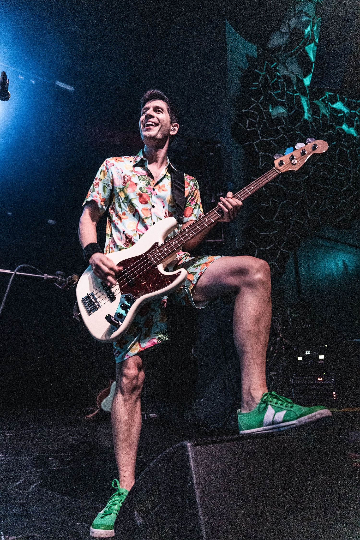 Bass player in floral shirt and green sneakers performs energetically on stage.