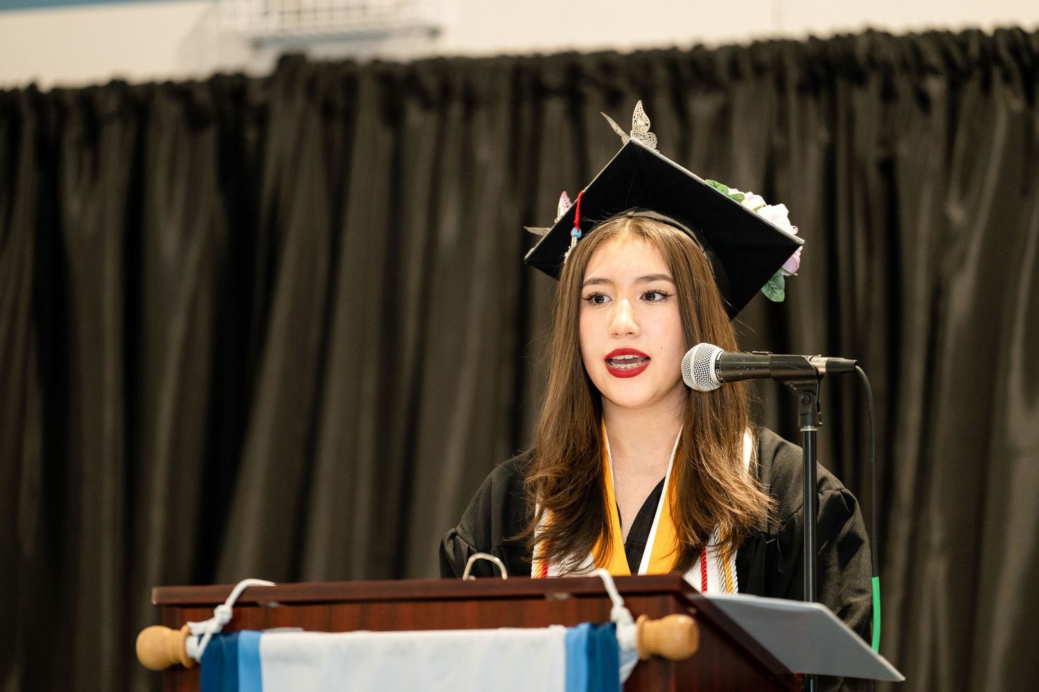Graduate in black cap and gown delivering speech at podium during commencement ceremony.