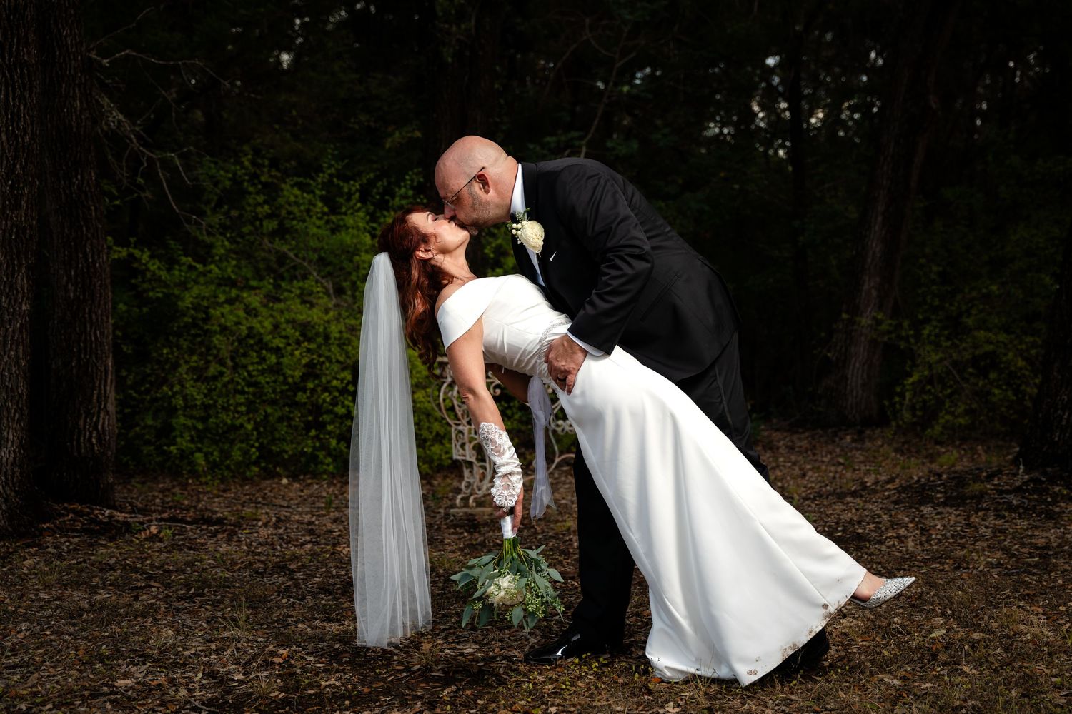 Romantic dip pose between couple in wedding attire against forest backdrop.