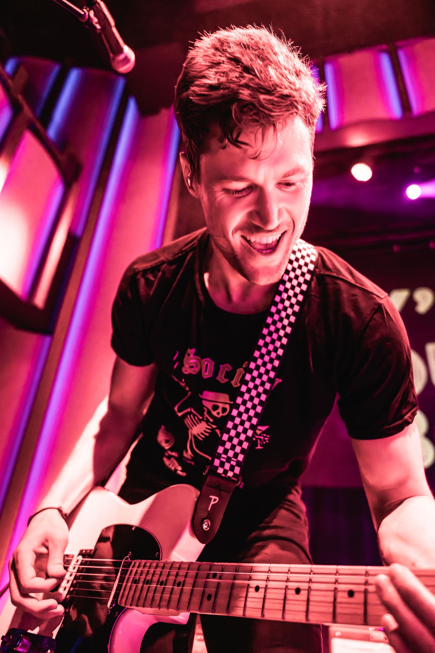 Musician playing guitar with joyful expression under red stage lighting.