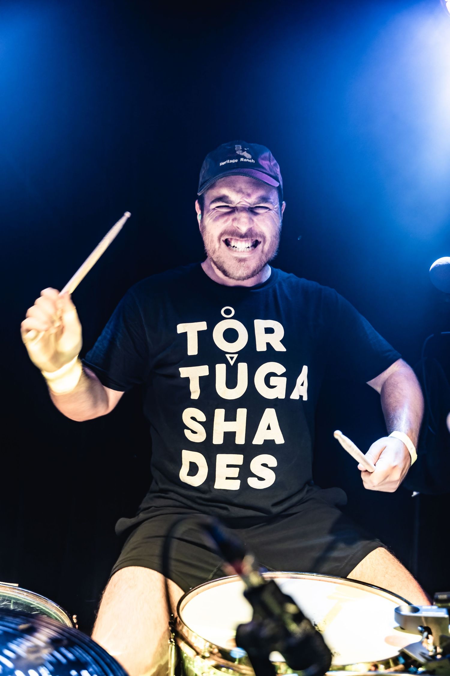 Drummer wearing a black t-shirt with text performs enthusiastically behind a drum kit under blue stage lighting.