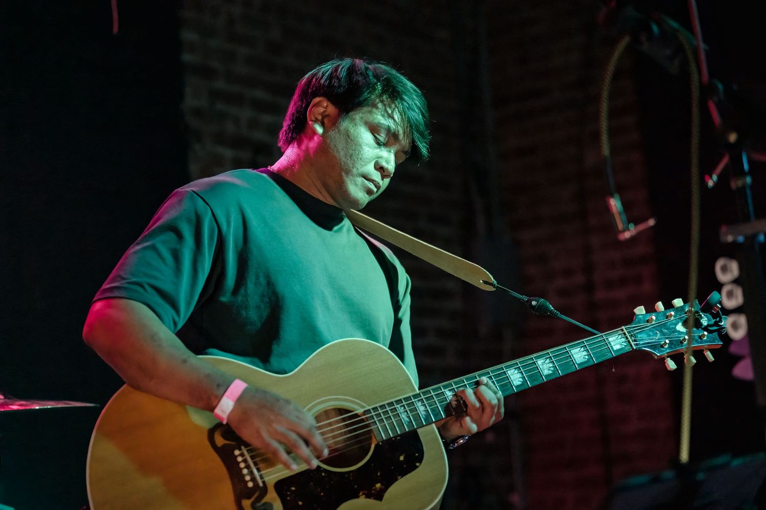 Acoustic guitarist performs on brick-walled stage with dramatic red and blue lighting effects.