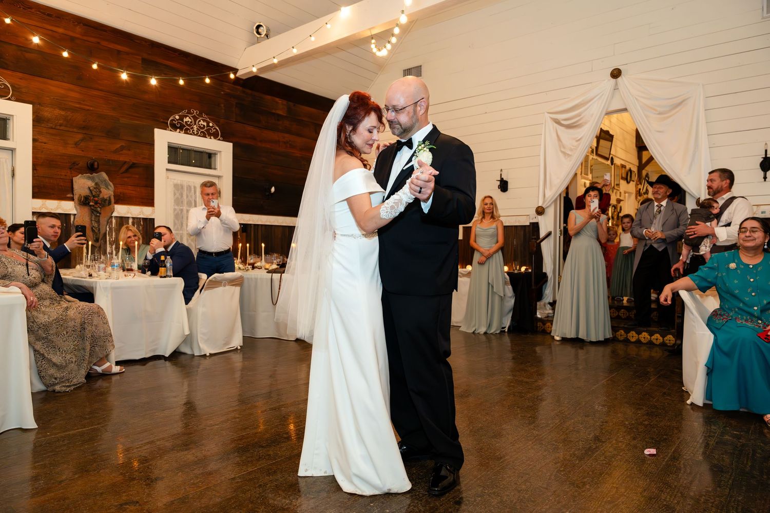 A couple dances surrounded by guests at their wedding reception with string lights overhead.