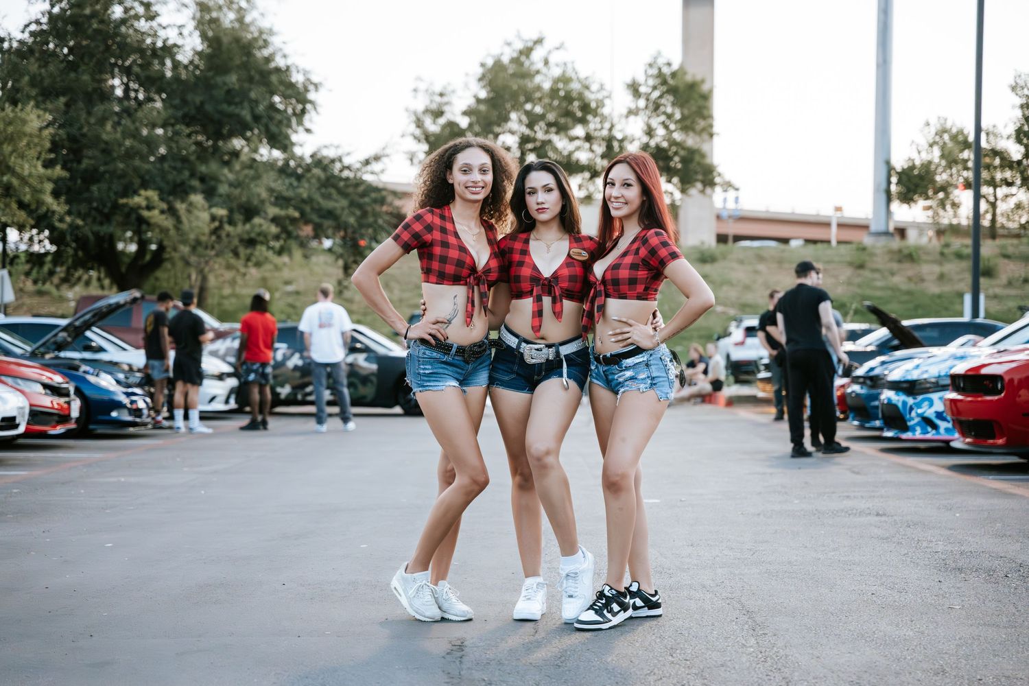 A car meet gathering with people posing in matching outfits in a parking lot with vehicles in the background.