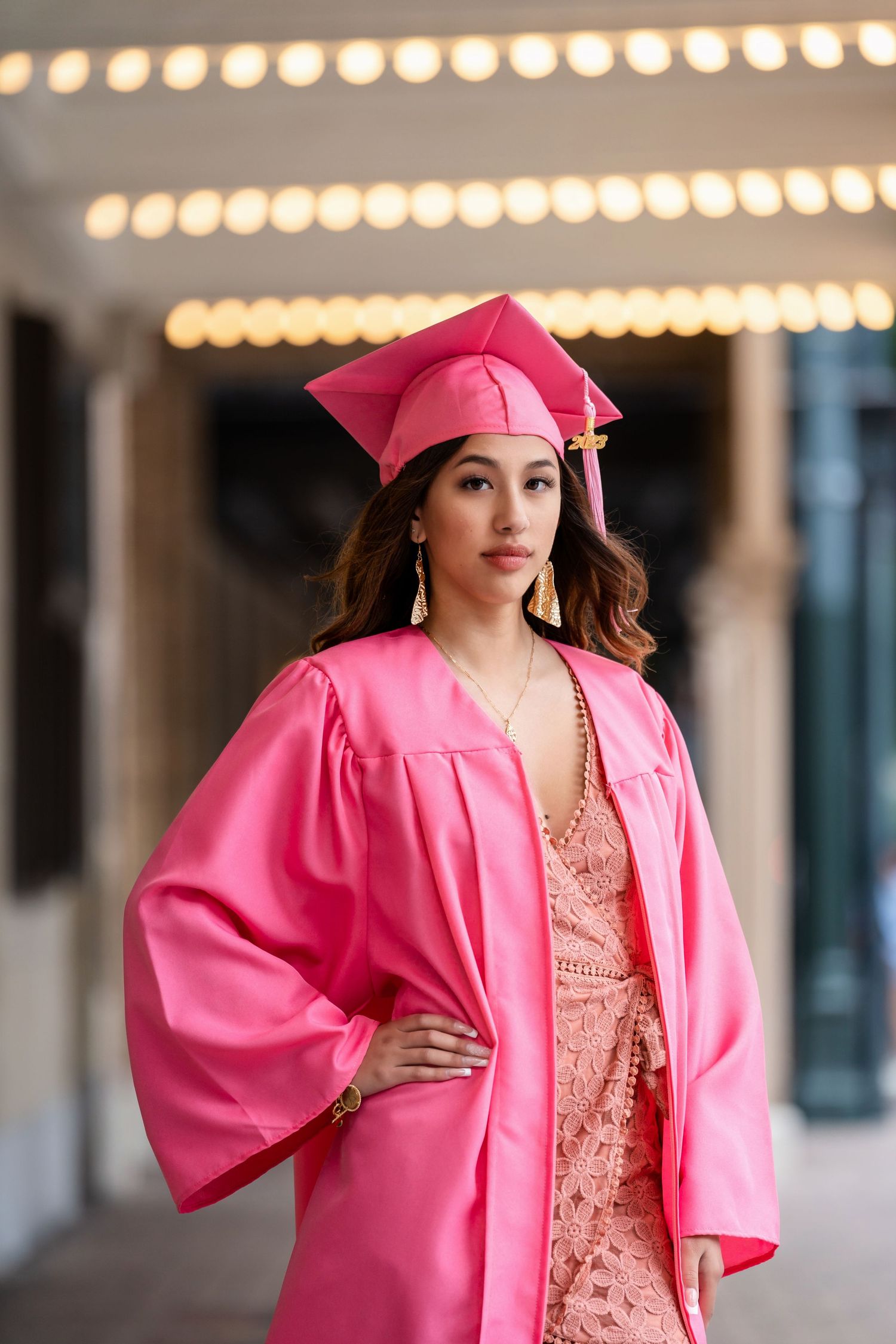 Graduate striking pose in pink graduation gown and lace dress under ambient lighting.
