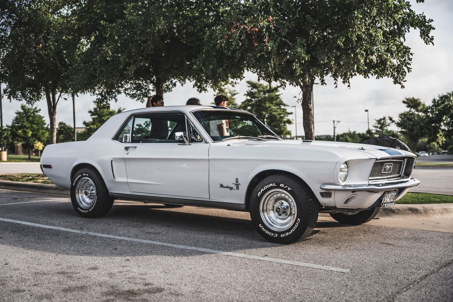 Classic white Ford Mustang coupe parked under trees in a parking lot.