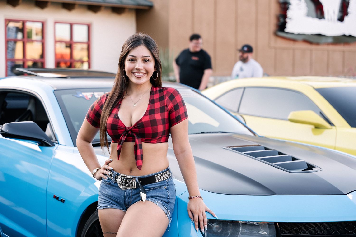 Person in red plaid crop top and denim shorts poses beside a blue sports car at a car meet.
