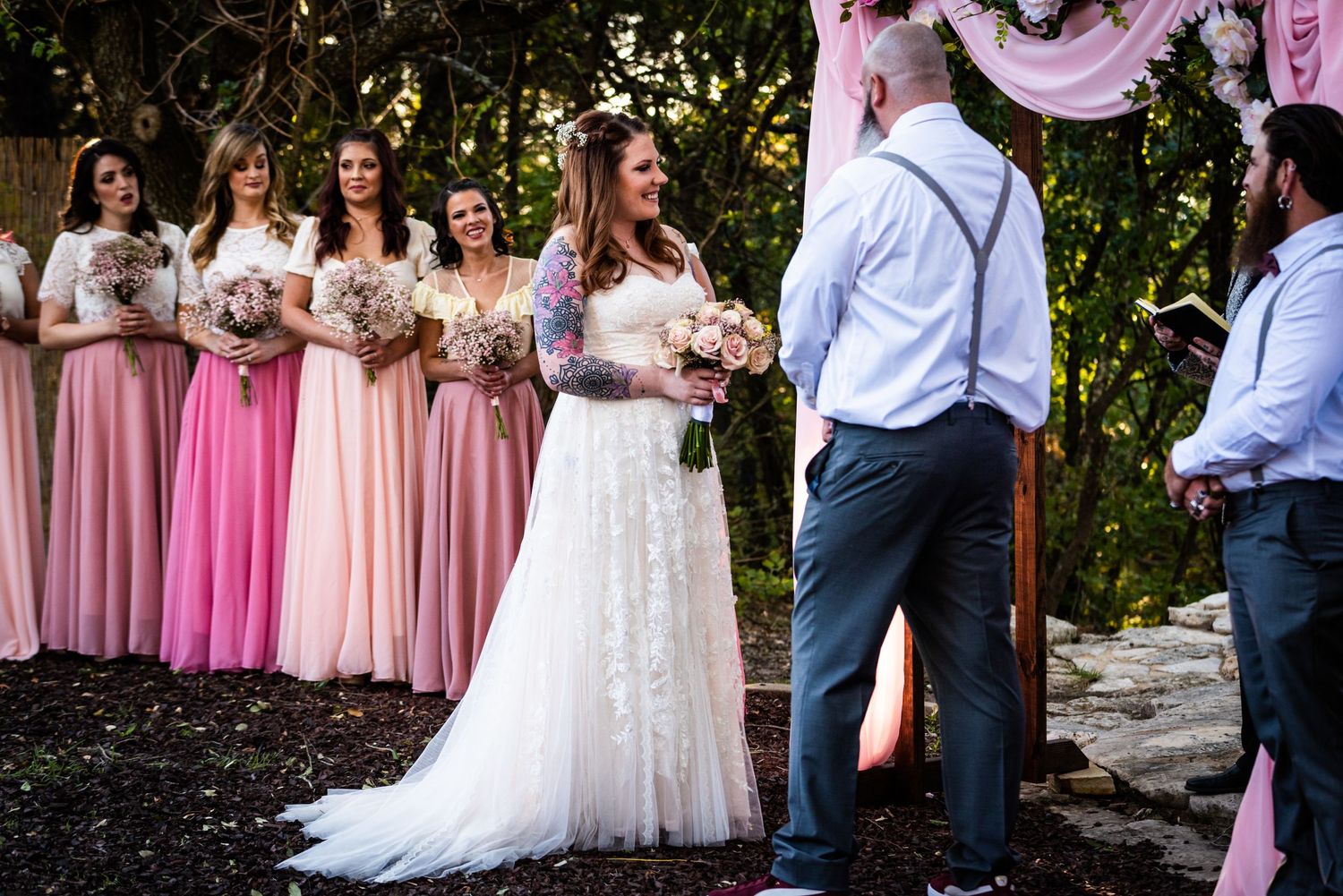 Wedding party stands during ceremony with bridesmaids in gradient pink dresses against garden backdrop.
