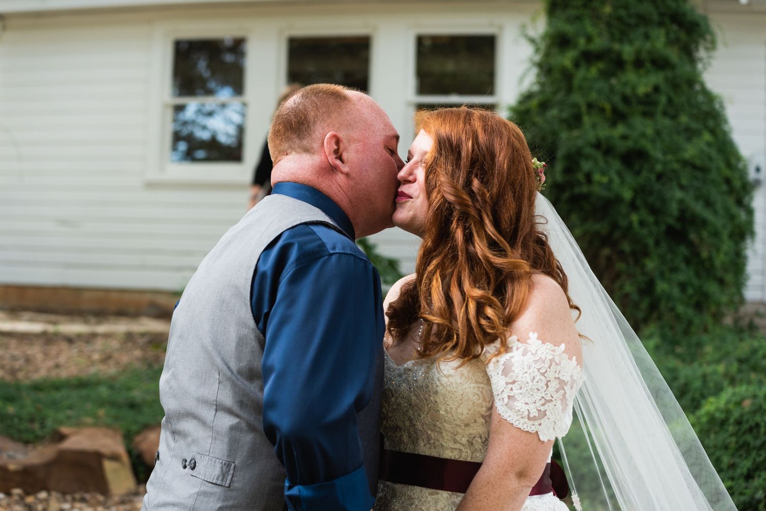 A romantic wedding kiss between newlyweds outdoors with a white house backdrop.
