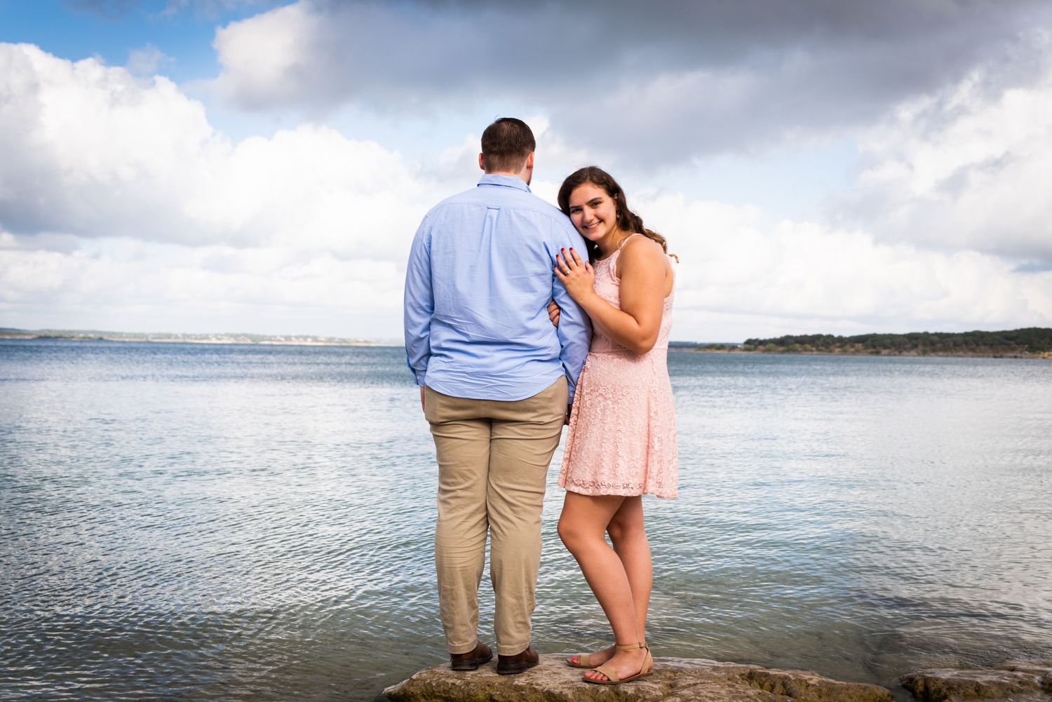 A couple stands together at the water's edge with cloudy skies above.