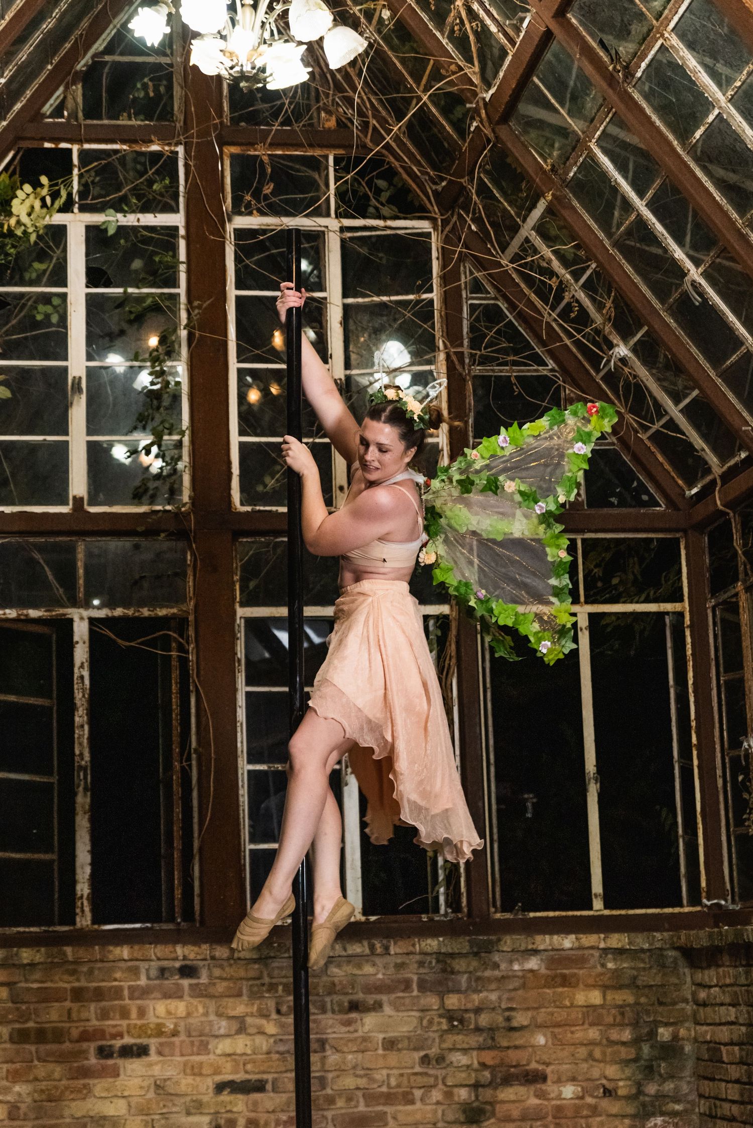 Dancer in peach dress performs on pole in rustic greenhouse setting.