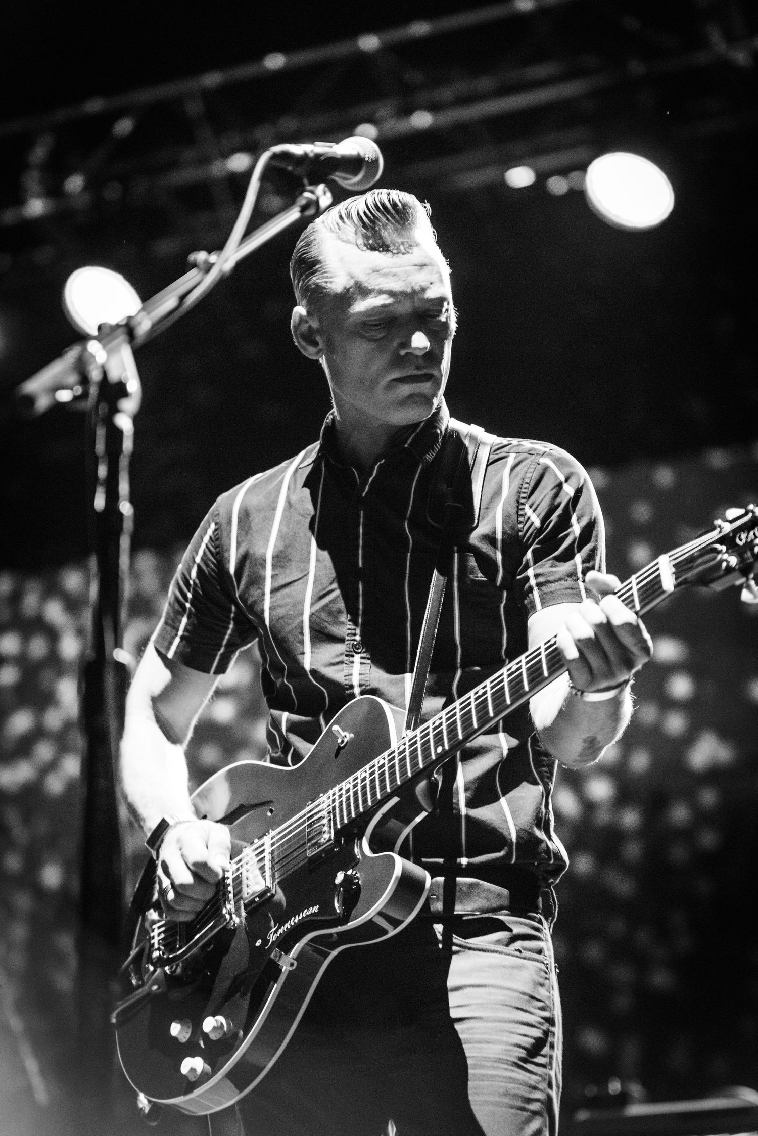 A guitarist in a striped shirt plays an electric guitar on a dimly lit stage with spotlights in the background.