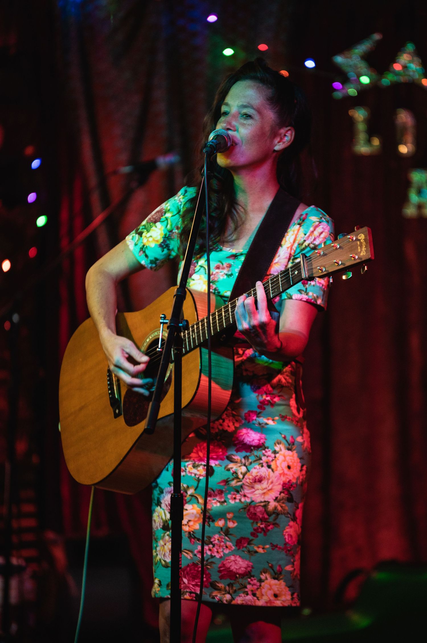 Musician in floral dress plays acoustic guitar during live performance at dimly lit venue.
