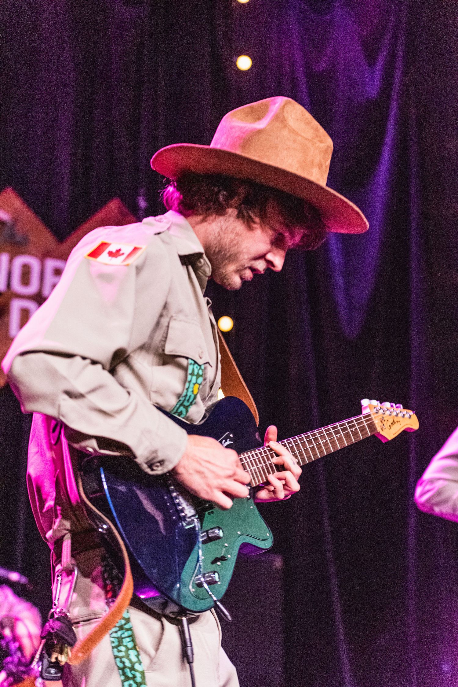 Guitarist in tan uniform and cowboy hat plays electric guitar under purple stage lights.