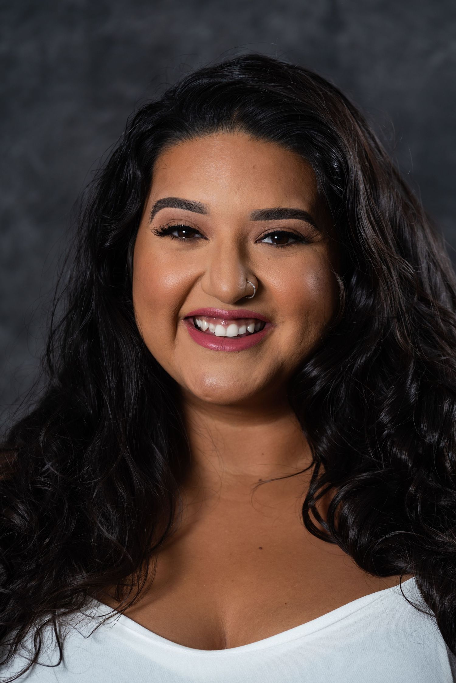 Professional portrait of a person with long dark curly hair wearing a white top against a dark background.