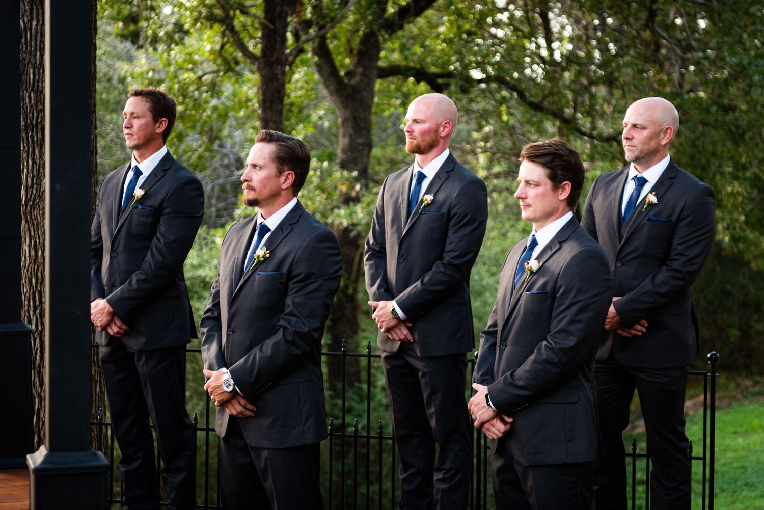 Groomsmen in dark suits stand in a line during an outdoor wedding ceremony.