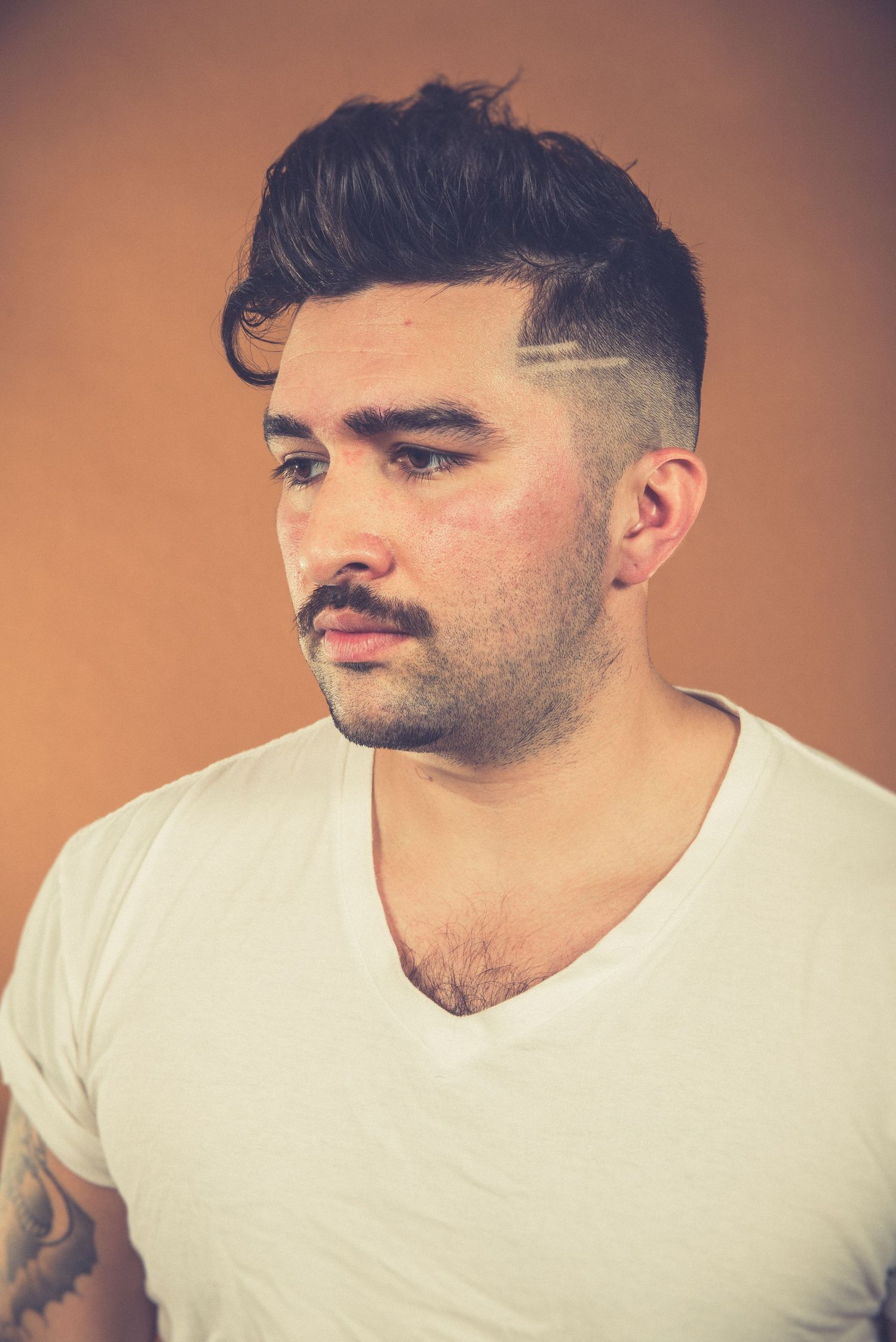 Portrait of someone in white v-neck t-shirt with styled hair against orange background.