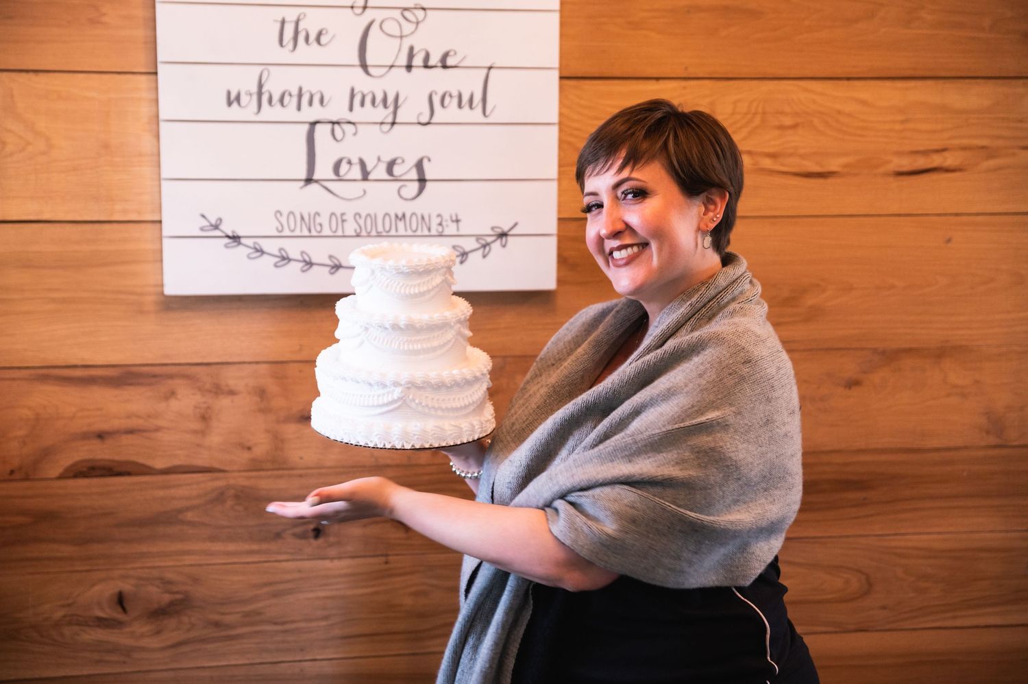 Person holding a white wedding cake in front of a wooden wall with text decor.