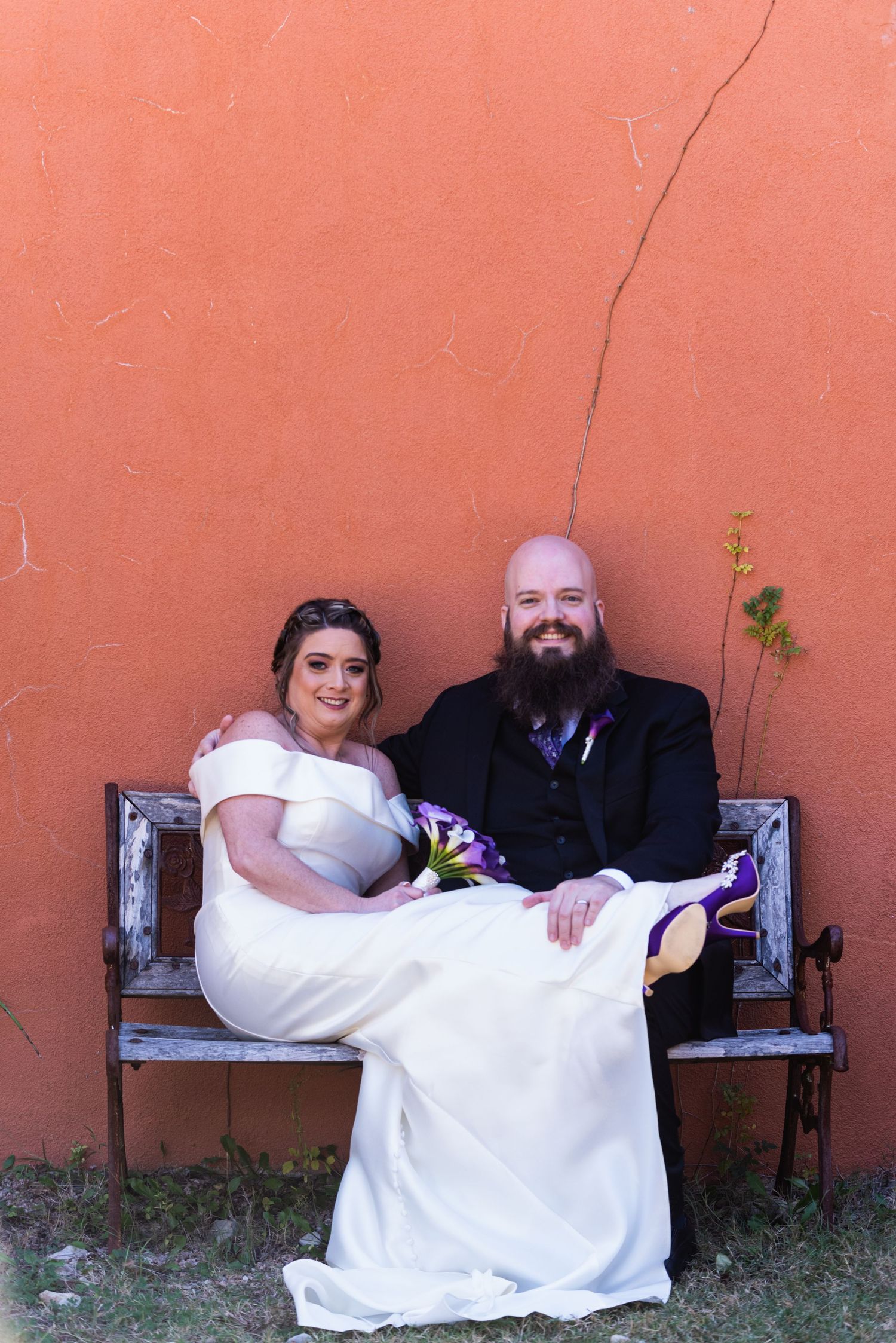A couple sits on a rustic bench against a coral colored wall.