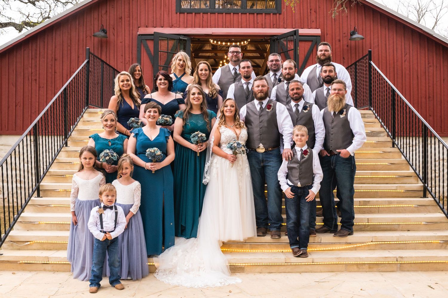 A wedding party poses on steps of a red barn with bridesmaids in teal dresses and groomsmen in vests.