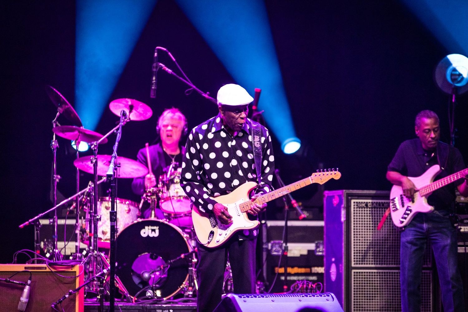 Band performs live with guitarist in polka dot shirt center stage under blue spotlights.