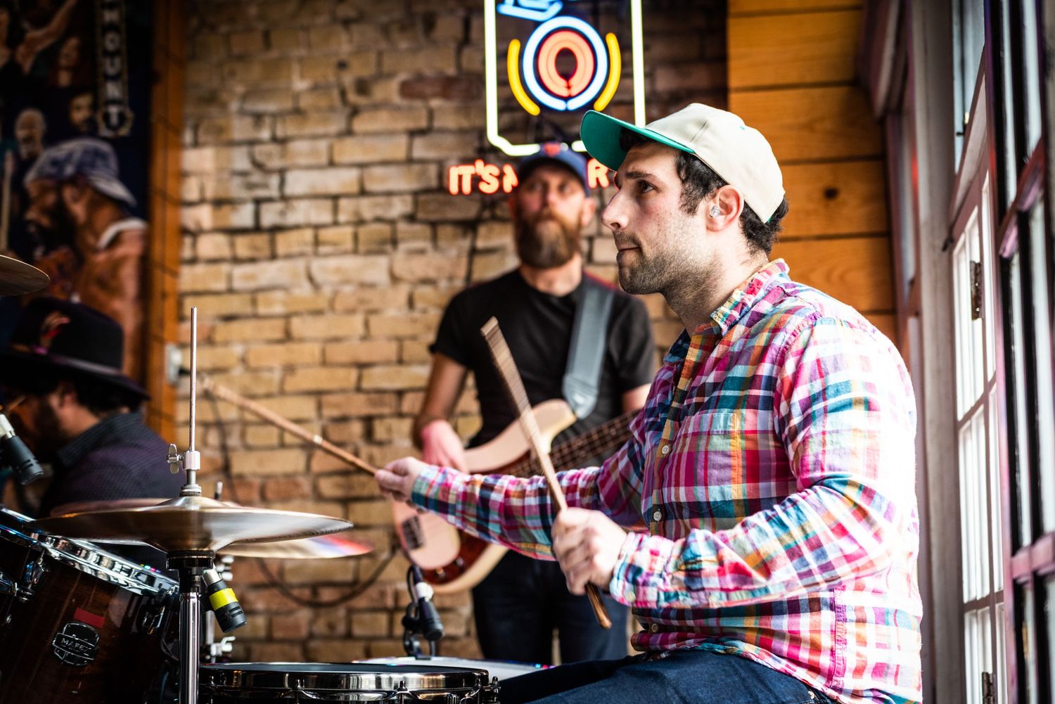Drummer performing with band in front of exposed brick wall with natural lighting.