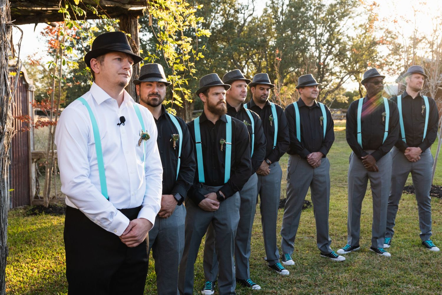 Groomsmen line up outdoors wearing matching turquoise suspenders and grey pants.