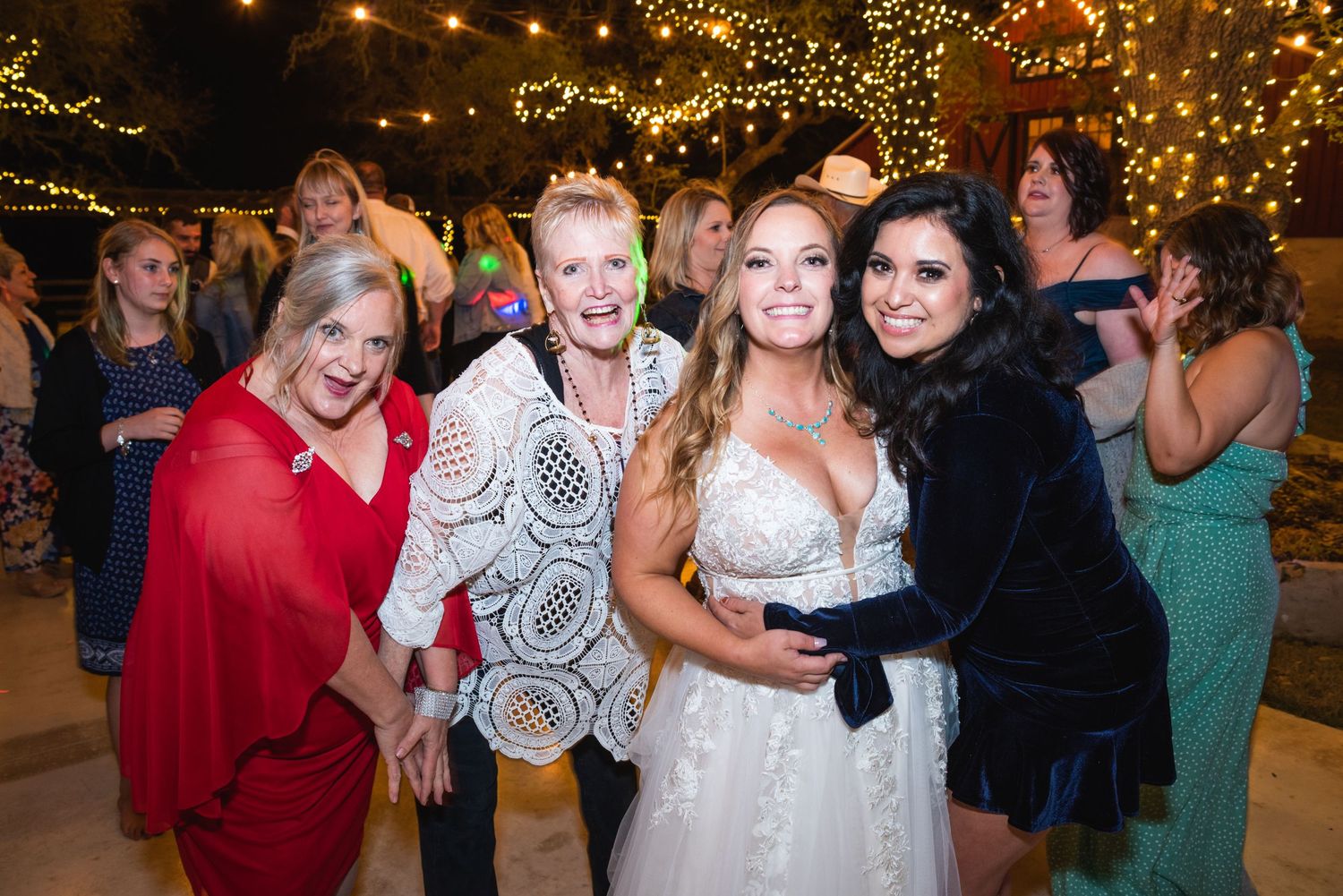 Wedding guests gather around the bride for a celebratory group photo under string lights.