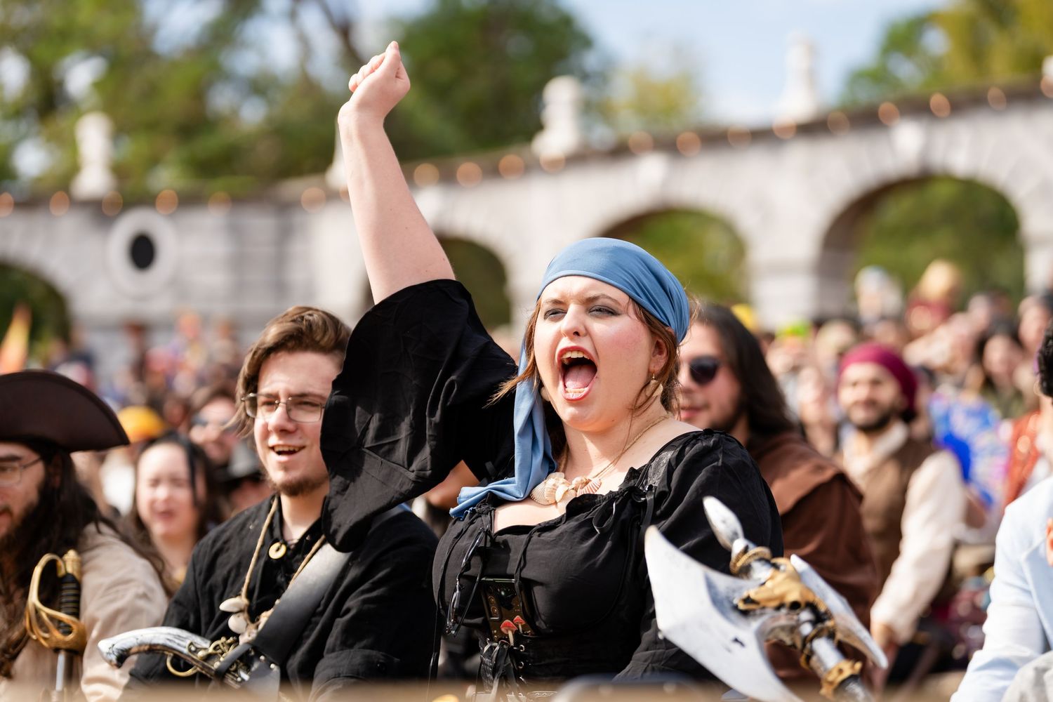 Excited concert crowd cheers and raises hands during an outdoor music festival performance on a sunny day.